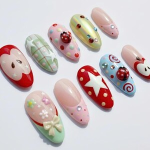 May include: A collection of ten colorful, decorated artificial nails. Designs include apples, ladybugs, stars, and floral patterns. Colors include red, pink, green, and blue, with various embellishments like rhinestones and bows.
