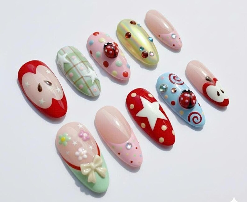 May include: A collection of ten colorful, decorated artificial nails. Designs include apples, ladybugs, stars, and floral patterns. Colors include red, pink, green, and blue, with various embellishments like rhinestones and bows.