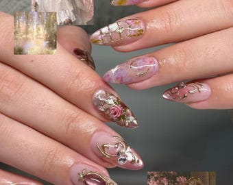 Puede incluir: Uñas en forma de almendra con una mezcla de diseños florales de acuarela en rosa y morado, detalles de filigrana dorada y dijes metálicos incrustados. La manicura incluye acabados brillantes, intrincados relieves dorados y cristales.