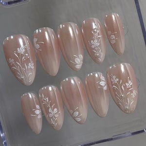 May include: A set of ten almond-shaped false nails. Each nail is a gradient of light pink to white, with a glossy finish. Delicate white floral designs with silver accents adorn each nail, creating an elegant and feminine aesthetic.