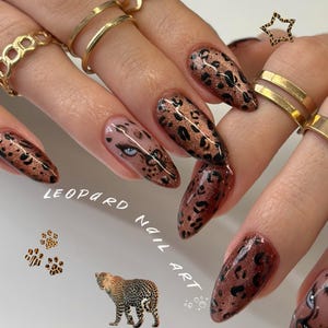 Puede incluir: Uñas en forma de almendra con una base de purpurina color cobre y manchas de leopardo negras. Una uña muestra una ilustración detallada de un ojo. El texto LEOPARD NAIL ART es visible junto a un gráfico de leopardo y huellas.