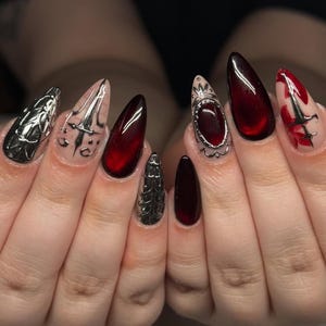 May include: A set of long, pointed fingernails with a dark red and silver design. Some nails are solid red, while others feature silver embellishments and intricate patterns. The nails are well-manicured and have a glossy finish.