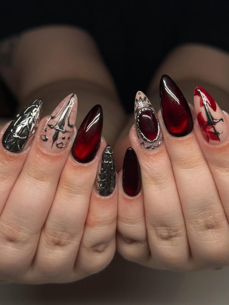 May include: A set of long, pointed fingernails with a dark red and silver design. Some nails are solid red, while others feature silver embellishments and intricate patterns. The nails are well-manicured and have a glossy finish.
