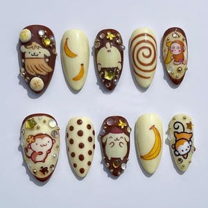 May include: A collection of ten artificial nails, each with a unique design. The nails showcase cartoon characters, bananas, and polka dots. The colour palette includes brown, cream, and gold, with rhinestones and beads. The nails are oval-shaped.