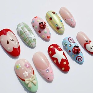 May include: A collection of ten artificial nails with various designs. The nails feature patterns like polka dots, stars, and ladybugs, and are painted in colors such as red, pink, green, and blue. Some nails have 3D embellishments.