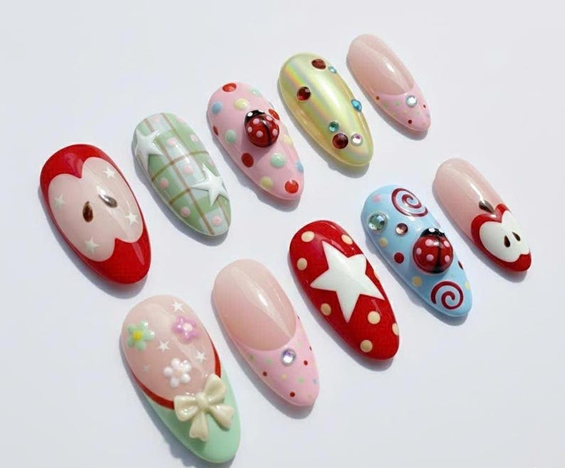 May include: A collection of ten artificial nails with various designs. The nails feature patterns like polka dots, stars, and ladybugs, and are painted in colors such as red, pink, green, and blue. Some nails have 3D embellishments.