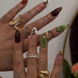 May include: A hand displays a variety of long, almond-shaped acrylic nails with unique designs. Some nails feature a tortoiseshell pattern, while others have green, white, and brown accents. Gold rings adorn the fingers, including a band of clear stones and a chunky, textured ring.