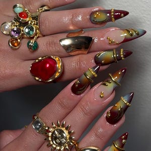 May include: Stiletto-shaped nails featuring a gradient transition from deep burgundy to yellow and clear tips, adorned with gold metal rings and studs. Several gold rings are worn on the fingers, including designs with colorful cabochons, a large red pearl, and a sun motif.