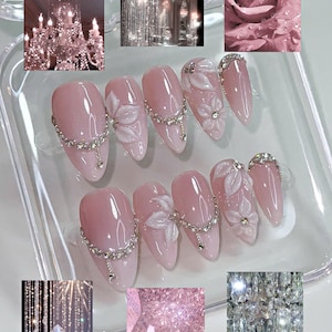 May include: A set of ten almond-shaped press-on nails in a glossy light pink finish. The nails feature three-dimensional white floral accents, embedded clear rhinestones, and delicate silver-toned chain details across the surface.