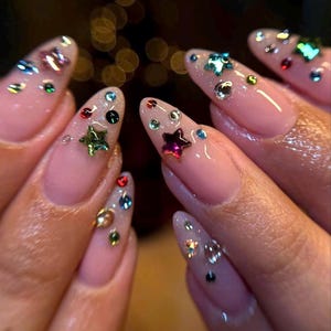 Puede incluir: Primer plano de uñas manicuradas con una base rosa claro. Cada uña está adornada con una variedad de pedrería colorida y adornos en forma de estrella, creando un efecto brillante. Las uñas son largas y almendradas.