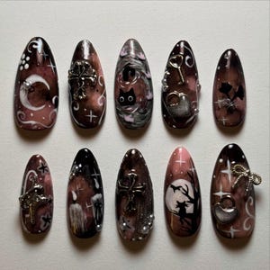 May include: A set of ten almond-shaped, dark brown and pink nail designs. Each nail features unique gothic-themed art, including moons, crosses, cats, keys, and heart-shaped locks. Some nails have silver embellishments.