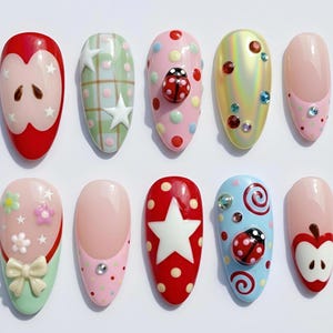 May include: A collection of ten artificial nails with various designs. The designs include an apple, stars, ladybugs, and floral patterns. Colors include red, pink, green, and blue, with decorative gems and bows.