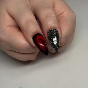 May include: Two long, pointed fingernails are visible. One nail is a gradient of red to black. The other nail is silver with a detailed, textured design. The nails are on a light background.