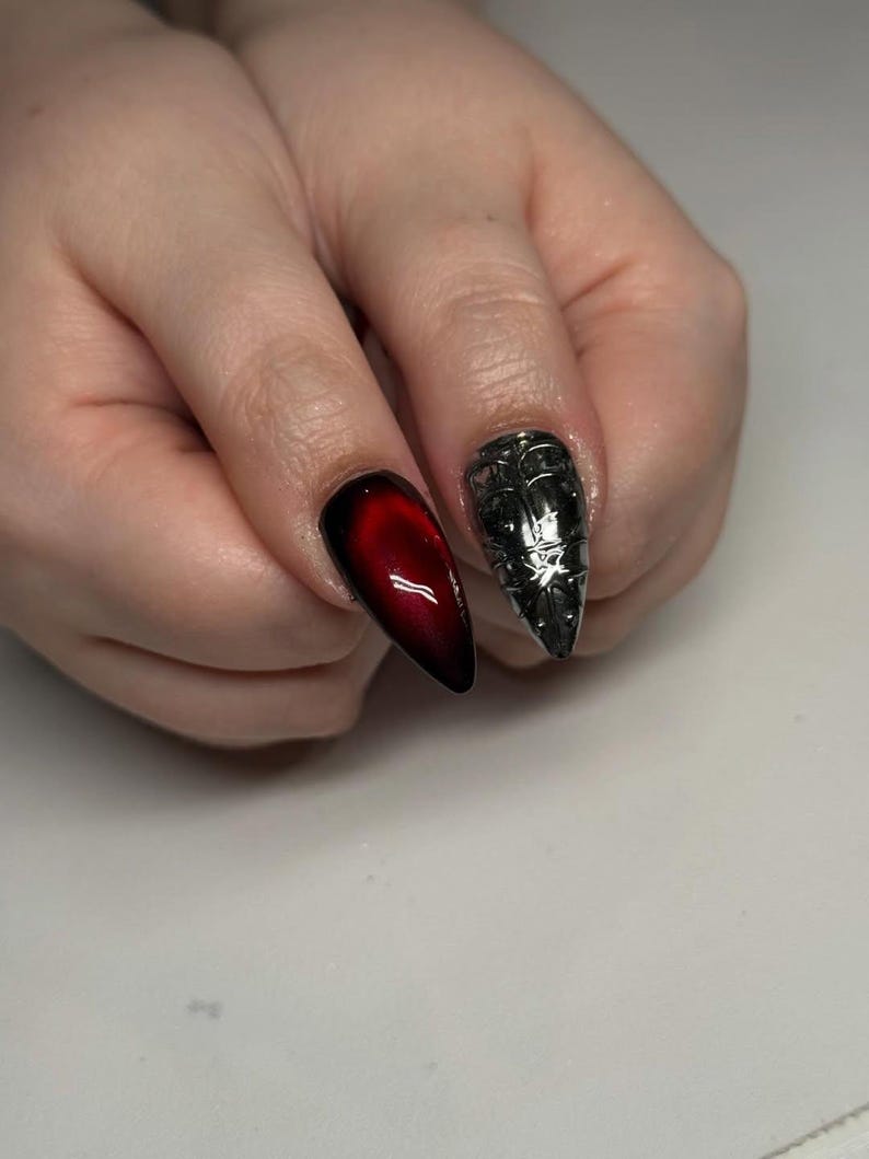May include: Two long, pointed fingernails are visible. One nail is a gradient of red to black. The other nail is silver with a detailed, textured design. The nails are on a light background.