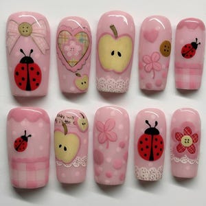 Puede incluir: Un conjunto de diez uñas postizas rosas con varios diseños. Los diseños incluyen mariquitas, manzanas, lazos, corazones y flores. Algunas uñas tienen un borde de encaje. Las uñas están decoradas con lunares y otros patrones bonitos.