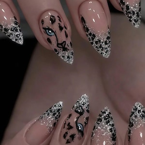 Puede incluir: Uñas en forma de almendra con base nude, puntas de purpurina plateada y patrones de estampado de leopardo negro. Una uña en cada mano muestra un ojo azul pintado a mano rodeado de manchas de leopardo negras, creando un diseño coherente.