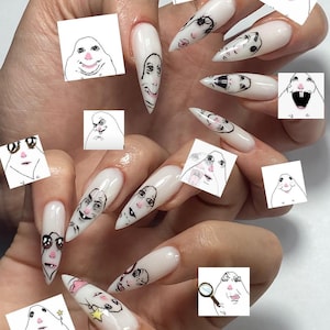 May include: Long, white acrylic nails with cartoon faces drawn on them. The nails are a glossy white and feature various cartoon faces in black ink. Small square images of cartoon faces are also scattered around the hand.