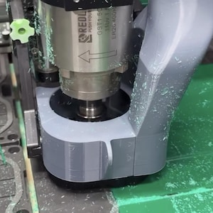 May include: Close-up of a CNC machine in operation, cutting a green plastic sheet. The machine has a silver spindle with the "REDL" logo and a gray housing. Green plastic shavings are visible around the cutting area.