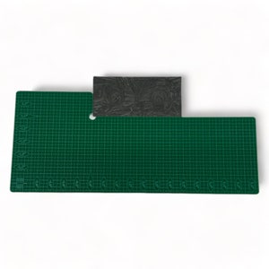 May include: A rectangular black rubber stamp featuring an intricate carved design of two circular motifs and foliage, positioned on a green self-healing cutting mat with white grid lines and imperial measurements in inches along the edges.