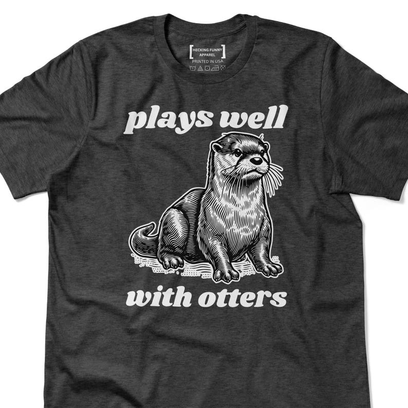 May include: Dark gray t-shirt with the text "plays well with otters" above and below a detailed illustration of an otter. The shirt has a crew neck and short sleeves. The brand name is printed on the upper left.