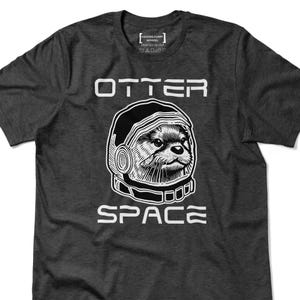 May include: Dark gray t-shirt with a graphic of an otter wearing a space helmet. The word "OTTER" is above the otter graphic, and the word "SPACE" is below. The shirt also has the text "HECKING FUNNY APPAREL PRINTED IN USA".