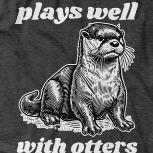 May include: Dark gray t-shirt featuring a detailed black and white illustration of an otter. The text "plays well" is above the otter, and "with otters" is below. The design has a vintage, engraved style.