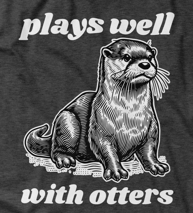 May include: Dark gray t-shirt featuring a detailed black and white illustration of an otter. The text "plays well" is above the otter, and "with otters" is below. The design has a vintage, engraved style.