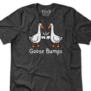 May include: Dark grey t-shirt with a cartoon design of two white geese with orange beaks and feet, facing each other. The text "Goose Bumps" is printed below the geese in white. The shirt has a crew neck.