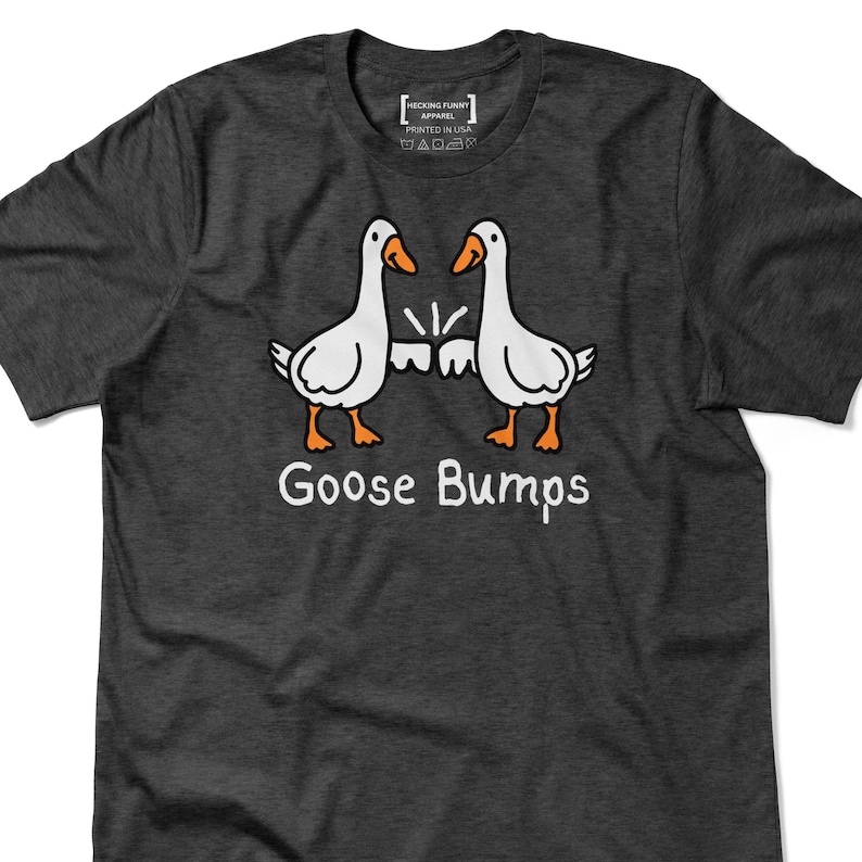 May include: Dark grey t-shirt with a cartoon design of two white geese with orange beaks and feet, facing each other. The text "Goose Bumps" is printed below the geese in white. The shirt has a crew neck.
