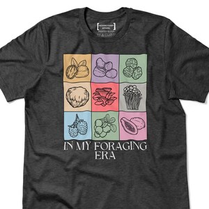 May include: Dark gray t-shirt featuring a grid of illustrated foraging items like mushrooms and berries. The text "IN MY FORAGING ERA" is below the grid. The shirt also has the text "HECKING FUNNY APPAREL PRINTED IN USA".