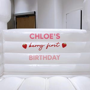May include: A white inflatable bounce house with a castle-like design. The text "CHLOE'S berry first BIRTHDAY" is printed in pink. Two white cone-shaped structures are on either side of the bounce house.