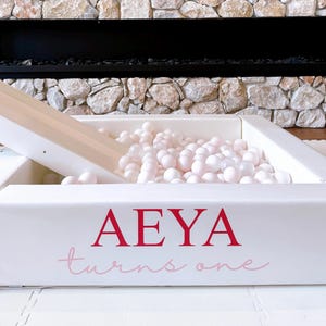 May include: A white ball pit with a slide, filled with light pink balls. The ball pit has the name "AEYA" and the phrase "turns one" printed on the side in red and pink lettering. The background features a stone wall.