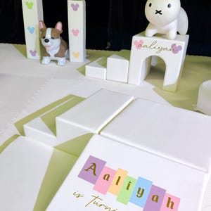 May include: White foam play set with a house-shaped arch, a corgi figurine, and a bunny figurine. The set includes blocks and a mat. The text "Aaliyah is turning Two" is displayed on a block.