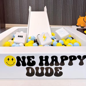 May include: A white ball pit with a small slide, filled with yellow, blue, and black balls. The pit has the text "ONE HAPPY DUDE" and a smiley face logo. There are three white cubes with different designs inside the pit.