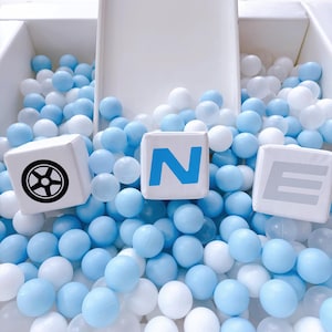 May include: A play area filled with light blue and white balls. Three white cubes with black and blue graphics are scattered among the balls. The cubes display a wheel, the letter "N", and the letter "E". A white slide is visible in the background.