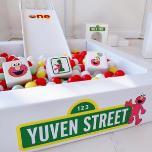 May include: A white play area with a slide and a ball pit filled with red, yellow, white, and green balls. The slide has the word "one" on it. The ball pit features blocks with Sesame Street characters and the words "Yuven Street" and "123 Yuven Street".