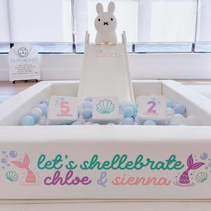 May include: A white ball pit with a small slide, filled with blue and white balls. A white bunny-shaped lamp sits atop the slide. The pit is decorated with mermaid-themed graphics and the text "Let's shellebrate Chloe & Sienna."