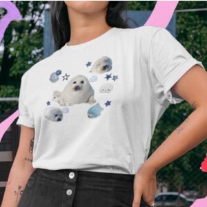 May include: White t-shirt featuring a playful design of cartoon seals in various poses. The seals are depicted in shades of white, gray, and blue, surrounded by stars and other aquatic elements. The shirt is a casual, short-sleeved style.