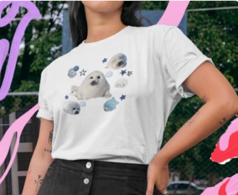 May include: White t-shirt featuring a playful design of cartoon seals in various poses. The seals are depicted in shades of white, gray, and blue, surrounded by stars and other aquatic elements. The shirt is a casual, short-sleeved style.