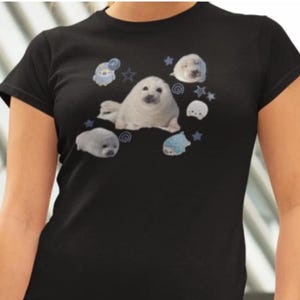 May include: Black t-shirt featuring a graphic of several white seals and blue stars and cartoon characters. The seals are in various poses, and the design is centered on the front of the shirt. The shirt has short sleeves and a crew neck.