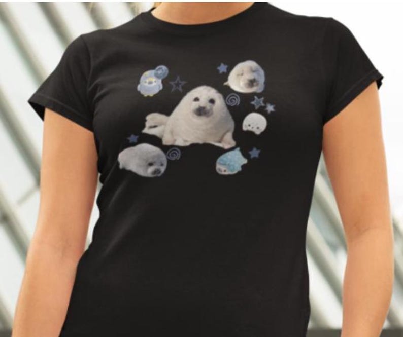 May include: Black t-shirt featuring a graphic of several white seals and blue stars and cartoon characters. The seals are in various poses, and the design is centered on the front of the shirt. The shirt has short sleeves and a crew neck.