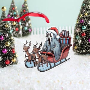 May include: A novelty Christmas ornament featuring a gray ghost character wearing a Santa hat, riding a red sleigh pulled by two reindeer. The sleigh is filled with wrapped gifts. The background includes miniature Christmas trees and a red ribbon.