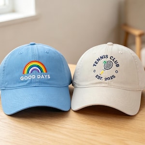 May include: A light blue baseball cap with an embroidered rainbow and the text "GOOD DAYS" and a cream-colored baseball cap with an embroidered tennis racket, a tennis ball, and the text "TENNIS CLUB EST. 2025" centered on the front panels.