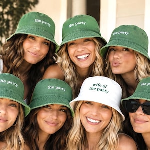Op de afbeelding: Een verzameling van zeven bucket hats, zes in een gewassen groene stof met de witte geborduurde tekst "the party" en één in witte stof met de donkergroene geborduurde tekst "wife of the party" gecentreerd op de voorzijde.