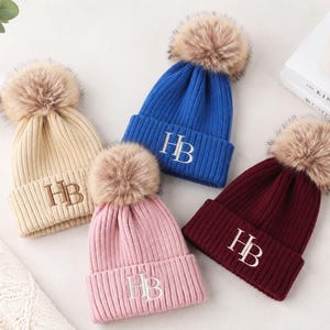 Puede incluir: Cuatro gorros de punto acanalado en beige, azul, rosa y burdeos, cada uno con un gran pompón de piel sintética en la parte superior y las letras HB bordadas en hilo blanco o marrón en el dobladillo doblado, formando un conjunto de invierno.