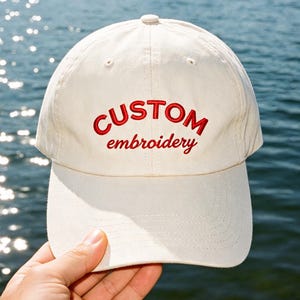 Op de afbeelding: Crèmekleurige baseballcap met de rode geborduurde tekst "CUSTOM embroidery". De cap heeft een gebogen klep en een knoop aan de bovenkant. De achtergrond is een watermassa.