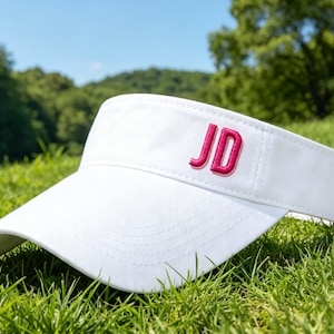 May include: A white visor with the embroidered letters "JD" in pink. The visor is resting on a bed of green grass, with a backdrop of trees and a blue sky.
