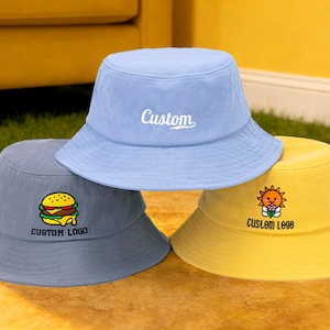 Könnte beinhalten: Drei Fischerhüte in Hellblau, Grau und Gelb. Der hellblaue Hut hat den weißen Schriftzug "Custom". Der graue Hut hat eine Burger-Grafik und "Custom Logo". Der gelbe Hut hat eine Sonnengrafik und "Custom Logo".