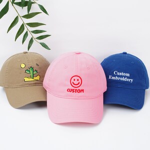 May include: Three baseball caps in a row. A tan cap with a cactus design, a pink cap with a smiling face and the word "CUSTOM", and a blue cap with "Custom Embroidery" in white text.