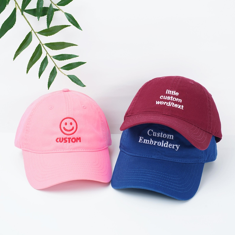 May include: Three baseball caps in pink, burgundy, and blue. The pink cap has a smiling face and the word "CUSTOM" embroidered in red. The burgundy cap says "little custom word/text" and the blue cap says "Custom Embroidery".